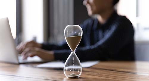 Woman working on computer with hour glass in foreground.